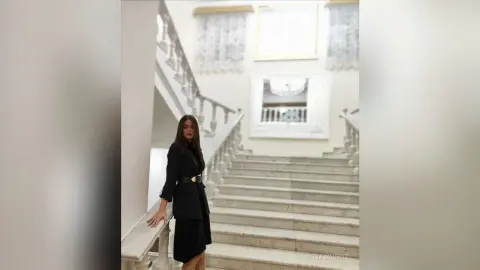 Elegant woman in a black blazer and skirt standing on a grand marble staircase in a bright, luxurious interior.