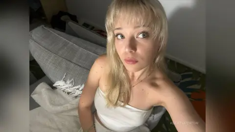 KeilaTouchton with blonde hair and bangs looks into the camera while sitting on a grey couch wearing a white tube top.