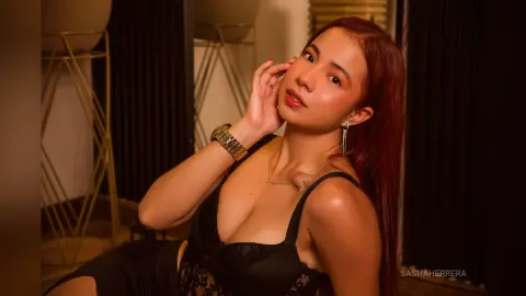 Glamorous woman in a black lace outfit posing with a confident expression in warm, moody indoor lighting.
