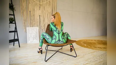 NataliiaSnake poses in a brown chair wearing a tight green snakeskin print bodysuit and black high heels, showing off her blonde hair and curves.