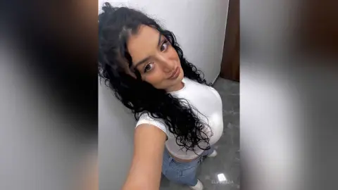 Smiling young woman with long curly dark hair wearing a white t-shirt in a bright indoor setting.