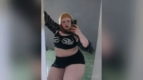 Redheaded woman in a black skeleton crop top and mini skirt taking a mirror selfie in a modern room.