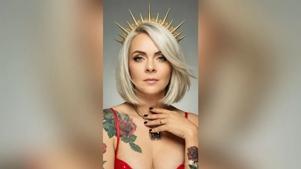 VickyAndrews — VickyAndrews wears a gold sun crown and red lingerie while posing with her silver hair and rose tattoos visible.
