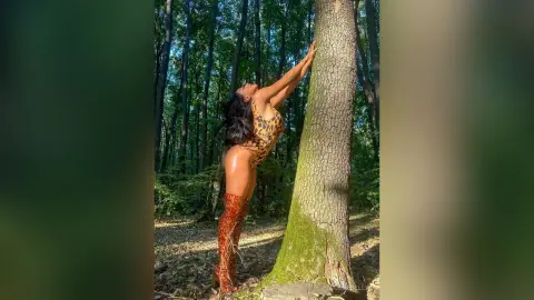NicoleBella stands naked in a forest, leaning her arms against a large tree trunk while wearing leopard print thigh-high boots.