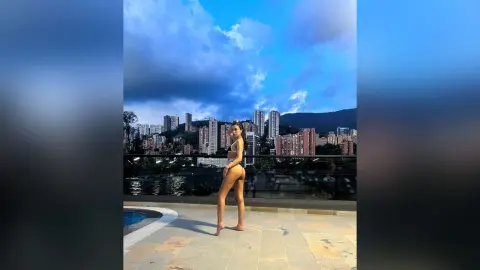 Slim woman in a white bikini posing on a luxury rooftop terrace overlooking a city skyline at dusk.