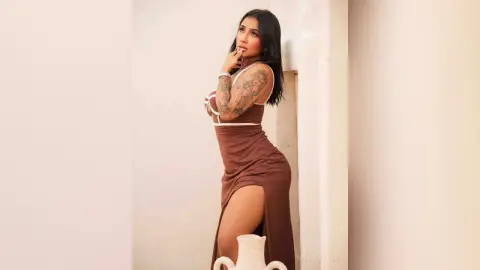 Tattooed woman in a brown sleeveless maxi dress posing elegantly against a minimalist white wall.