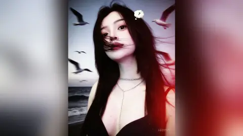 HedyIvy poses with long dark hair and a silver chain necklace, wearing a black top against a beach background with flying seagulls.