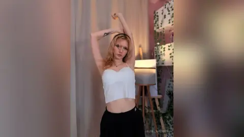 Blonde woman in a white crop top and black skirt posing gracefully in a bright, minimalist room.