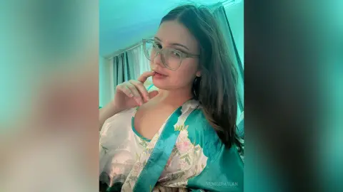 Young woman in glasses and a floral teal silk saree posing thoughtfully in a bright room.
