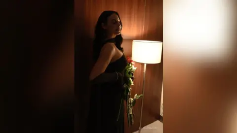 Elegant woman in a black evening gown holding red roses in a moody, dimly lit room.