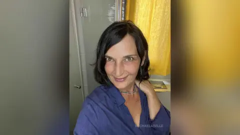 MichaelaBelle smiles while wearing a blue button-down shirt against a bathroom background with a yellow shower curtain.