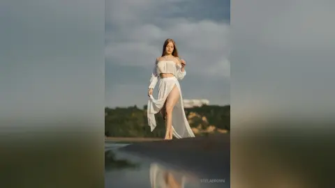StelaBrown stands on a sandy beach wearing a white off-the-shoulder top and a long, sheer white skirt that shows her bare legs.