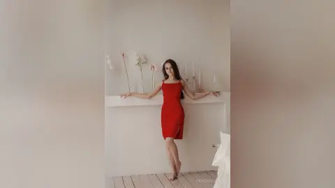 IngridBeckton stands in a white room wearing a tight red dress, leaning her arms against a white ledge with her long dark hair down.