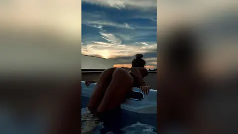 Woman in swimwear relaxing in a rooftop pool under a dramatic sunset sky.