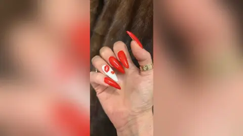 Close-up of long red almond-shaped nails with white lip art design on a hand wearing a gold ring.