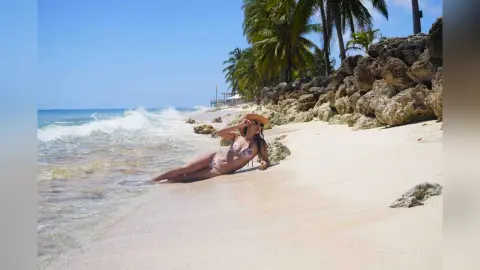 LucyMartin reclines on a sandy beach wearing a patterned bikini, with ocean waves and palm trees in the background.