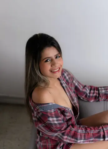 Smiling young woman in a red plaid shirt relaxing indoors against a neutral wall.