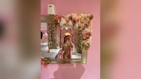 Woman in pink lingerie taking a mirror selfie in a dreamy room decorated with lush floral arrangements.