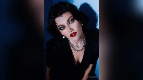 Glamorous woman with red lipstick and silver chain necklaces posing in moody blue studio lighting.