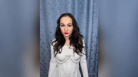 EllaVietor poses in a white long-sleeve dress with pearl trim against a blue floral curtain background.