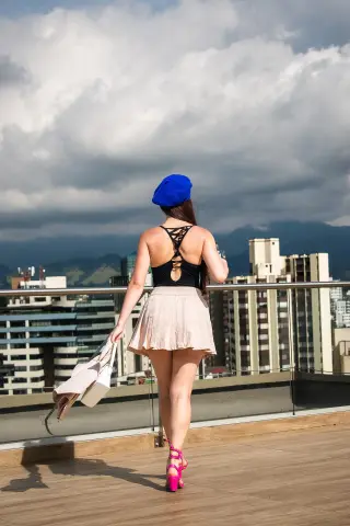 Woman in a black tank top and pink skirt walking on a rooftop balcony overlooking a city skyline under cloudy skies.
