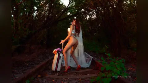 Elegant woman in white lingerie and veil standing by a suitcase in a moody, dark forest setting.