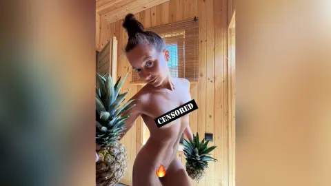 Nude woman with hair in a bun posing in a sunlit wooden room next to a pineapple.