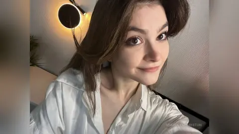 Smiling young woman with long brown hair wearing a white button-down shirt in a cozy, dimly lit room.