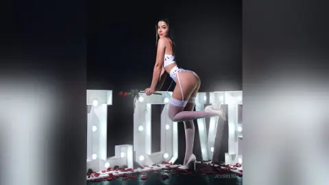 JessieLiu poses in white lace lingerie and white thigh-high stockings, standing next to large illuminated letters on a bed of red rose petals.