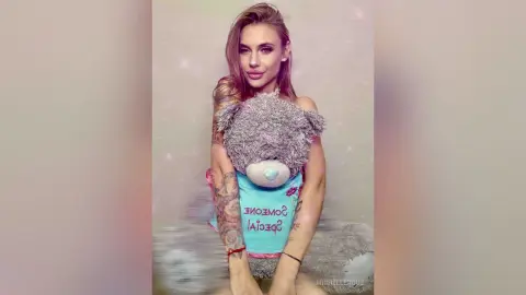Glamorous woman in a playful teddy bear graphic tank top posing against a dreamy pink background.