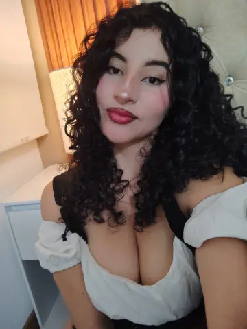 Young woman with curly dark hair wearing a white off-the-shoulder top in a cozy bedroom setting.
