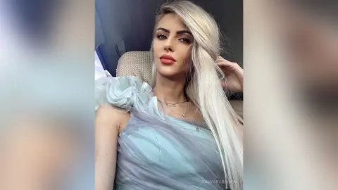 Blonde woman in a light blue ruffled dress posing with a glamorous look in a car interior.