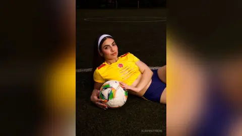 AlmaWhitmore lies on a dark grass field wearing a yellow short-sleeved shirt and blue shorts, holding a soccer ball near her stomach.