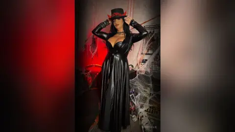 MissyJolie stands wearing a long black leather dress and a Halloween hat, posing with her hands up by her head.