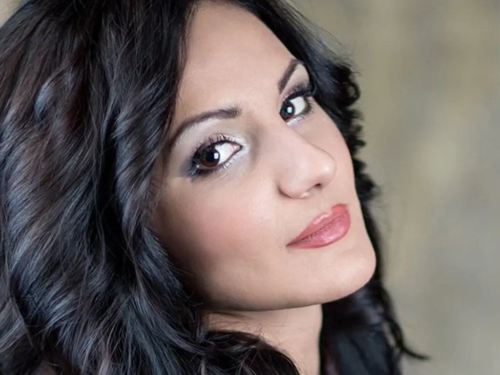 Close-up portrait of a smiling woman with wavy dark hair and glamorous makeup in soft natural lighting.