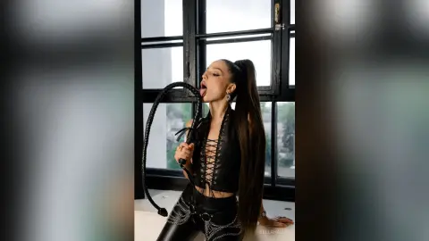 Playful woman in edgy black leather corset and pants holding a whip in a bright modern room.