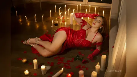 Glamorous woman in a red lace bodysuit reclining on a dark floor surrounded by glowing candles and rose petals.