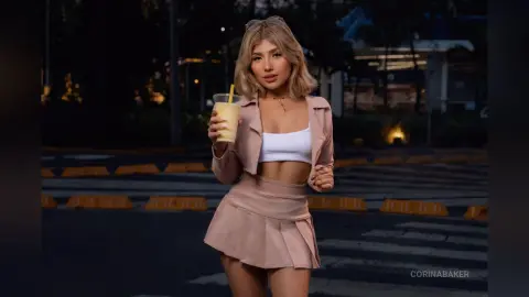 Stylish woman in a beige crop top and mini skirt holding an iced latte in a dark outdoor parking lot setting.