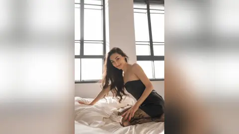 Smiling woman in a black strapless dress sitting on a white bed in a bright, minimalist sunlit room.