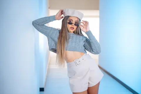 Stylish woman in a grey knit sweater and white shorts posing in a bright, minimalist hallway.