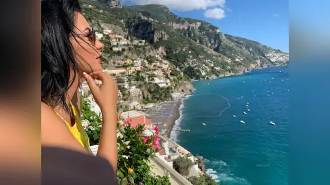 MissyJolie looks out over a coastal town and blue ocean while wearing a yellow top with her hand resting on her chin.
