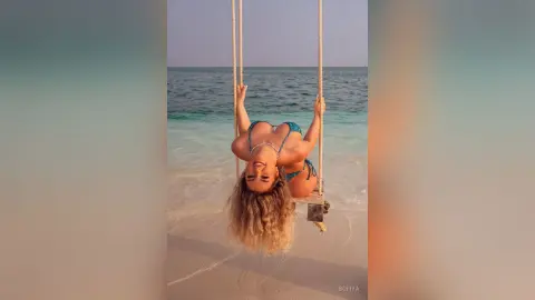 Cheerful blonde woman in a blue bikini relaxing on a beach swing over turquoise ocean waves at sunset.