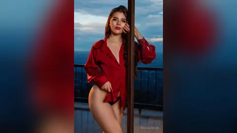 Glamorous woman in a red silk shirt posing on a balcony during a moody sunset.