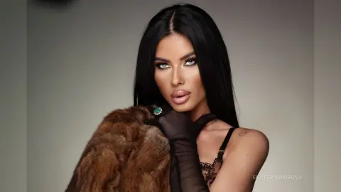 Glamorous woman with long black hair wearing a brown fur coat and black lace lingerie in a studio setting.