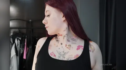 Red-haired woman with floral neck tattoos wearing a black tank top in a moody indoor setting.