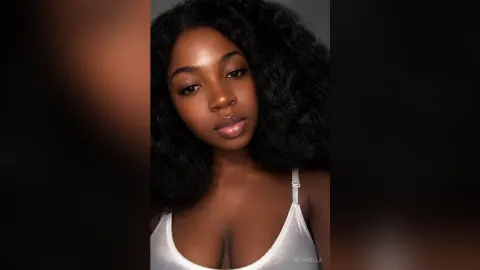Misskella — Misskella poses with long wavy black hair wearing a white tank top against a dark background.