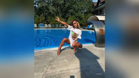 Stylish woman in a white oversized shirt and bikini posing by a sunny outdoor swimming pool.