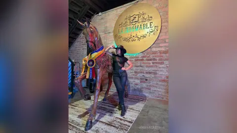 Stylish woman in black top and jeans posing next to a large horse statue in a rustic indoor setting.