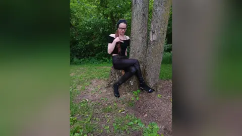 Woman in black casual outfit sitting on a tree stump in a lush green forest setting.
