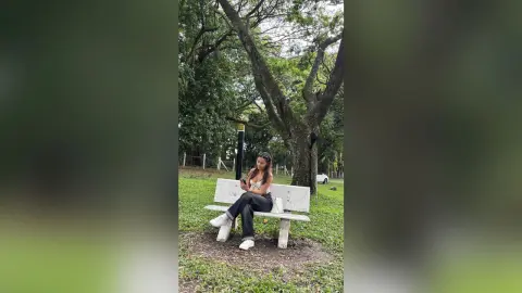 Young woman in a white crop top and black jeans sitting on a park bench under lush green trees.